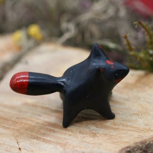 May include: A small black ceramic figurine of a fox with a red tip on its tail. The fox is standing on a wooden surface.
