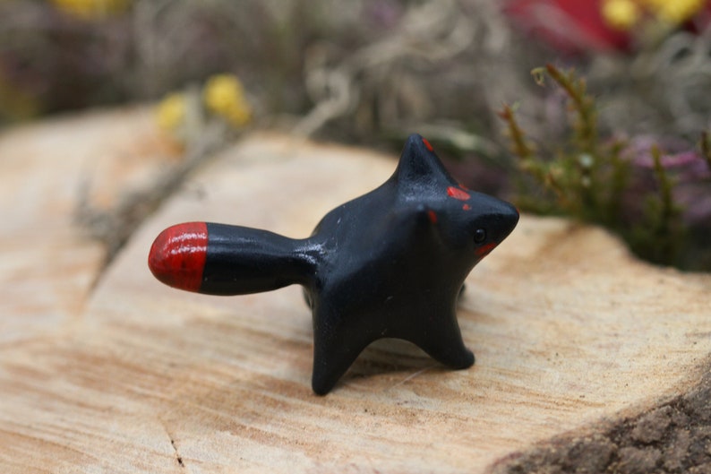 May include: A small black ceramic figurine of a fox with a red tip on its tail. The fox is standing on a wooden surface.