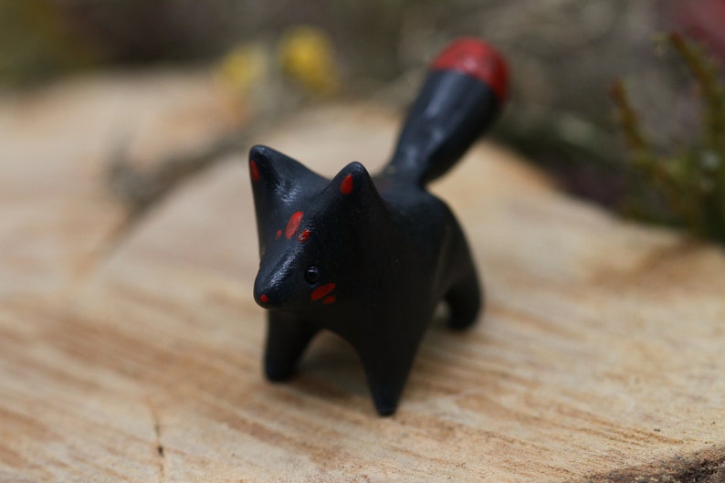 May include: A small black ceramic fox figurine with red accents on its ears, face, and tail. The figurine is standing on a wooden surface.