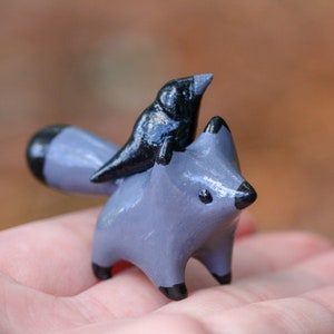 K&ouml;nnte beinhalten: Eine kleine graue Keramik-Fuchsfigur mit einem schwarzen Vogel, der auf seinem R&uuml;cken sitzt. Der Fuchs hat einen schwarzen Schwanz und schwarze Augen.