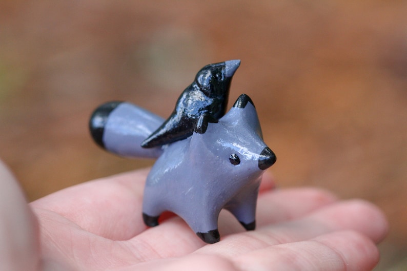 K&ouml;nnte beinhalten: Eine kleine graue Keramik-Fuchsfigur mit einem schwarzen Vogel, der auf seinem R&uuml;cken sitzt. Der Fuchs hat einen schwarzen Schwanz und schwarze Augen.