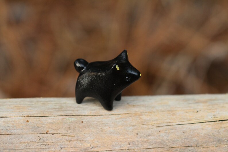 May include: A small black ceramic figurine of a dog with yellow eyes. The figurine is standing on a piece of wood.