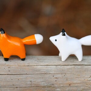 Pu&ograve; includere: Due piccole statuine di volpe in ceramica, una arancione e una bianca, con accenti neri sulle orecchie e le code. Le volpi sono in piedi su una superficie di legno.
