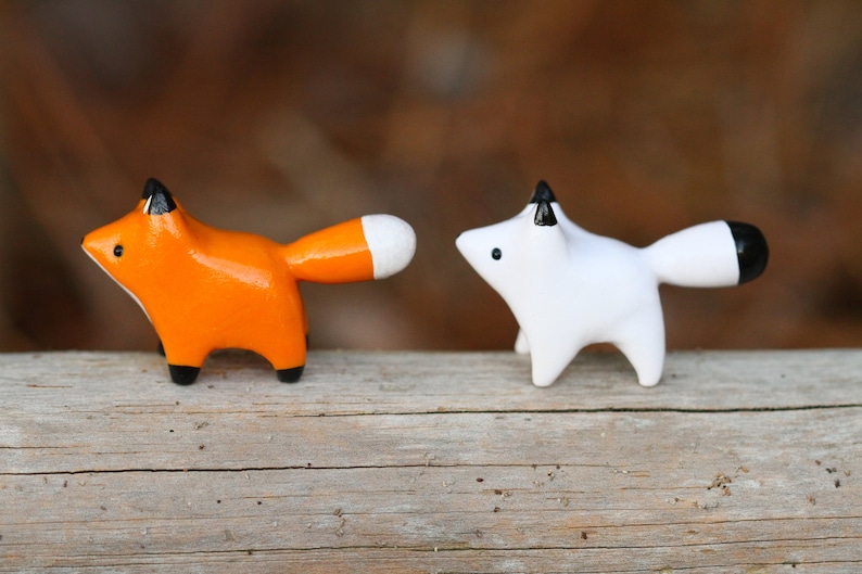 Pu&ograve; includere: Due piccole statuine di volpe in ceramica, una arancione e una bianca, con accenti neri sulle orecchie e le code. Le volpi sono in piedi su una superficie di legno.