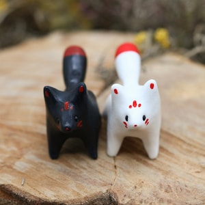 May include: Two ceramic fox figurines, one black with red accents and one white with red accents. The foxes are standing on a wooden surface.