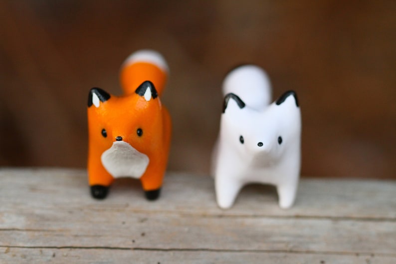 Pu&ograve; includere: Due statuine di volpe in ceramica, una arancione con accenti neri e un petto bianco, l'altra bianca con accenti neri. Entrambe le volpi sono in piedi su una superficie di legno.