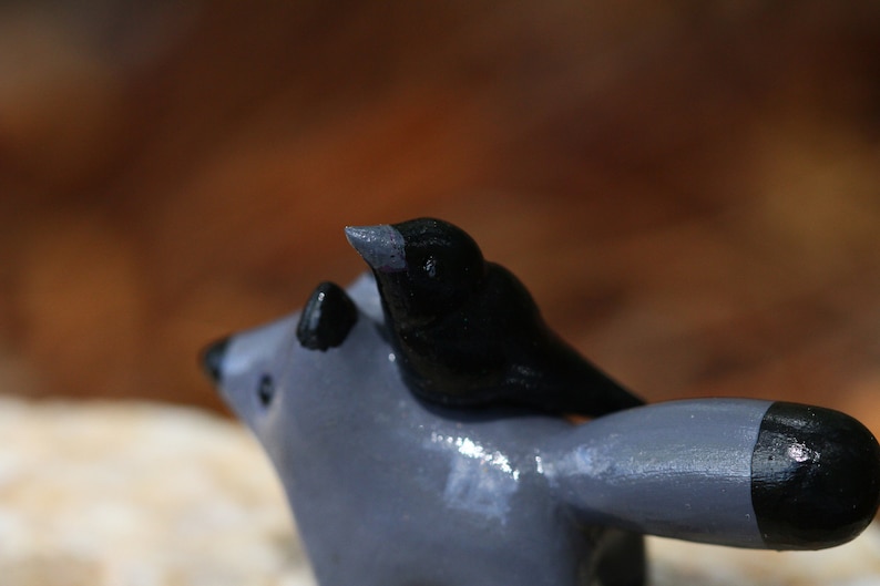 K&ouml;nnte beinhalten: Eine graue und schwarze Keramikfigur eines Waschb&auml;ren mit einem schwarzen Vogel auf dem R&uuml;cken.