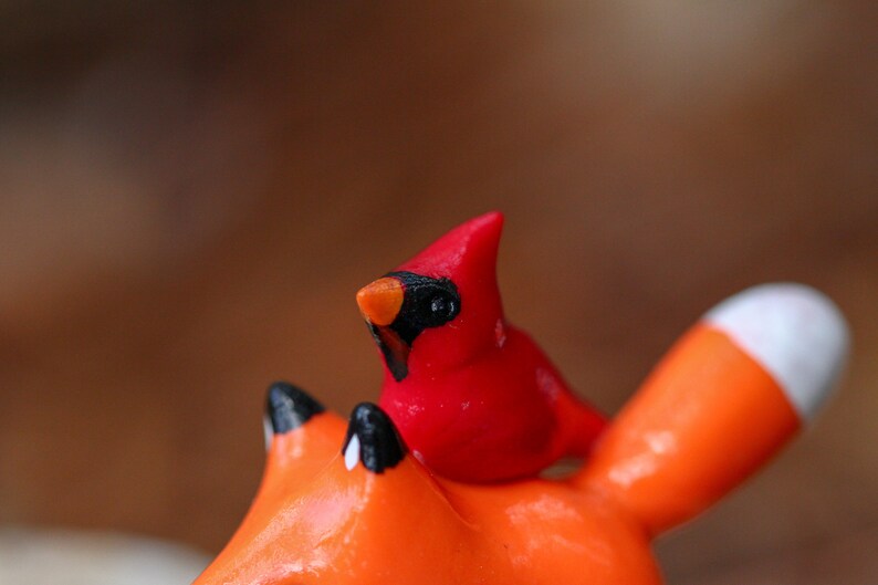 K&ouml;nnte beinhalten: Eine kleine rote Kardinalsfigur, die auf einer gr&ouml;&szlig;eren orangefarbenen Fuchsfigur sitzt. Beide Figuren sind aus bemalter Keramik.