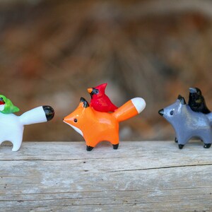 K&ouml;nnte beinhalten: Drei Keramik-Tierfiguren mit V&ouml;geln auf ihren R&uuml;cken. Die erste ist ein wei&szlig;er Fuchs mit einem gr&uuml;nen Kolibri, die zweite ist ein orangefarbener Fuchs mit einem roten Kardinal und die dritte ist ein grauer Fuchs mit einem schwarzen Vogel.