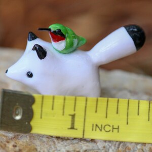 K&ouml;nnte beinhalten: Eine wei&szlig;e Keramik-Fuchsfigur mit schwarzer Schwanzspitze und einem gr&uuml;n-roten Kolibri, der auf ihrem R&uuml;cken sitzt. Die Figur ist etwa 2,5 cm hoch und steht auf einem gelben Ma&szlig;band.