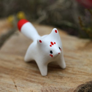 May include: White ceramic fox figurine with red accents on the tail and face. The fox is standing on a wooden surface.