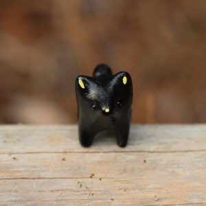 May include: A small black ceramic figurine of a fox with yellow eyes. The fox is standing on a wooden surface.