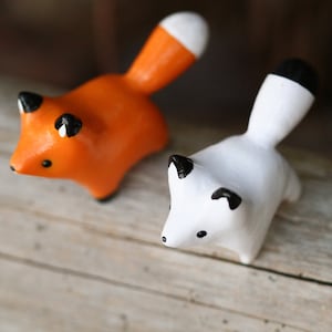 Pu&ograve; includere: Due piccole figurine di volpe in ceramica, una arancione e una bianca, con occhi e nasi neri. Le volpi sono in piedi su una superficie di legno.