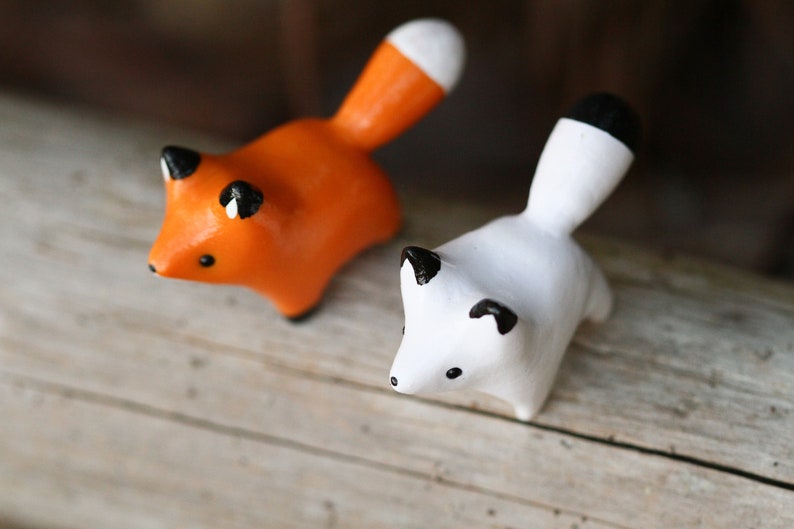 Pu&ograve; includere: Due piccole figurine di volpe in ceramica, una arancione e una bianca, con occhi e nasi neri. Le volpi sono in piedi su una superficie di legno.