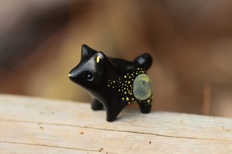 May include: A small black ceramic figurine of a wolf with yellow accents. The wolf has a round, yellow moon on its back with yellow dots scattered across its body.