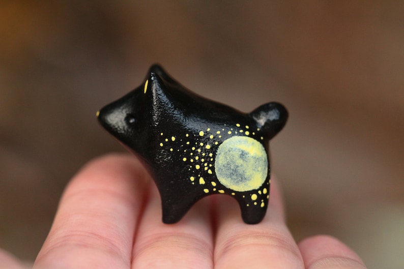 May include: A small black ceramic animal figurine with yellow paint details. The figurine has a large yellow circle on its back, resembling a moon, and smaller yellow dots scattered across its body.