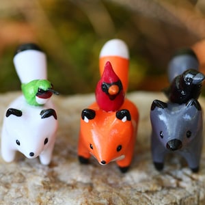 K&ouml;nnte beinhalten: Vier kleine Keramik-Tierfiguren mit V&ouml;geln auf dem Kopf. Die Tiere sind ein wei&szlig;er Fuchs mit einem Kolibri, ein orangefarbener Fuchs mit einem Kardinal, ein grauer Fuchs mit einer schwarzen Kr&auml;he und ein wei&szlig;er Fuchs mit einem Kolibri.