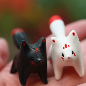 May include: Two small ceramic fox figurines, one black with red accents and one white with red accents. The foxes are standing on a hand.