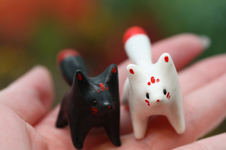 May include: Two small ceramic fox figurines, one black with red accents and one white with red accents. The foxes are standing on a hand.