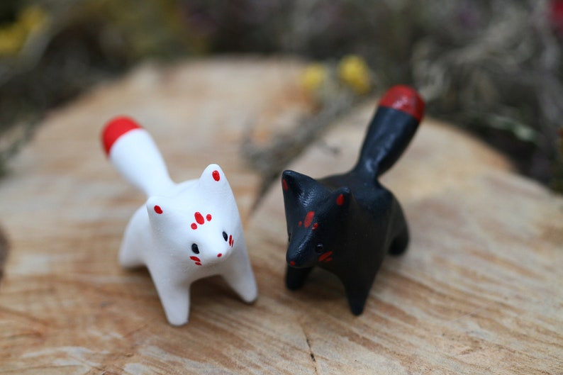 May include: Two small ceramic fox figurines, one white with red accents and the other black with red accents. The figurines are standing on a wooden surface.
