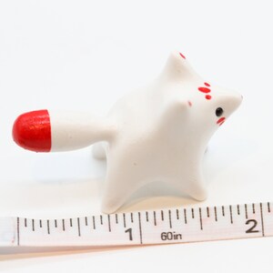 May include: White ceramic figurine of a fox with red accents on the tail and face. The figurine is about 2 inches long and is sitting on a white surface next to a ruler.
