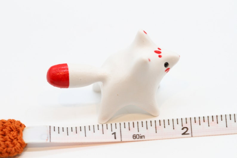 May include: White ceramic figurine of a fox with red accents on the tail and face. The figurine is about 2 inches long and is sitting on a white surface next to a ruler.
