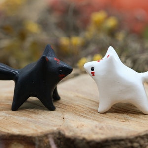 May include: Two small ceramic fox figurines, one black with a red tip tail and the other white with a red tip tail, facing each other on a wooden surface.