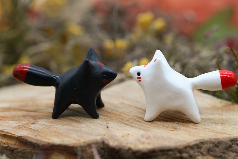 May include: Two small ceramic fox figurines, one black with a red tip tail and the other white with a red tip tail, facing each other on a wooden surface.