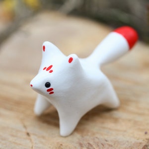 May include: A small white ceramic fox figurine with red markings on its face and tail. The figurine is standing on a wooden surface.
