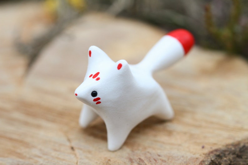 May include: A small white ceramic fox figurine with red markings on its face and tail. The figurine is standing on a wooden surface.