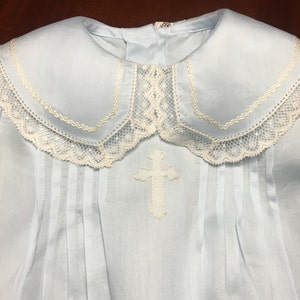 May include: A light blue christening gown with a white cross applique. The gown features a Peter Pan collar trimmed with lace. Vertical pleats run down the front of the garment, adding texture and detail to the design.