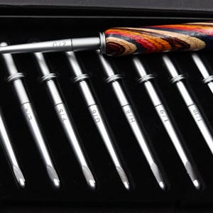 May include: A set of eight metal crochet hooks with silver shafts and size markings ranging from D/3 to K/10.5. One hook features a multicolored, layered wood handle with red, orange, yellow, blue, and dark brown stripes, while the others are plain.