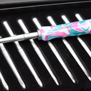 May include: A set of silver metal crochet hooks with a single hook featuring a marbled teal and pink acrylic handle. The hooks are arranged in a black case, with the metal shafts clearly marked with size letters including C/2, D/3, E/4, F/5, and G/6.