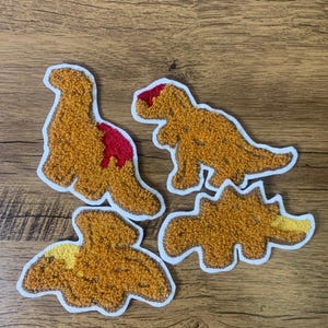 May include: Four felt dinosaur patches in brown, yellow, and red. The patches are outlined in white and have a fuzzy texture.