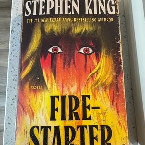May include: A vintage paperback book titled "Firestarter" by Stephen King. The cover features a fiery illustration with a woman's face and the title in bold, black letters. The book is a novel and is from the #1 New York Times bestselling author.