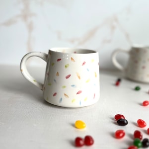 May include: White ceramic mug with a handle, featuring a scattered pattern of colorful painted leaves. The mug is sitting on a white surface with colorful jelly beans scattered around it.