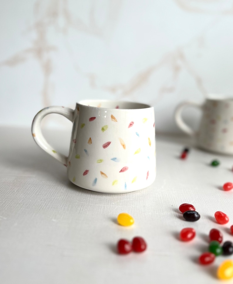 May include: White ceramic mug with a handle, featuring a scattered pattern of colorful painted leaves. The mug is sitting on a white surface with colorful jelly beans scattered around it.