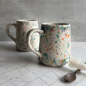 May include: Two ceramic mugs with a speckled design in blue, green, orange, and yellow on a white background. Each mug has a curved handle. A wooden spoon and a white decorative element are also visible.