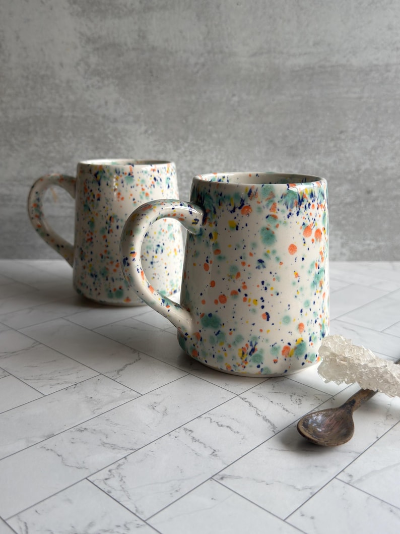 May include: Two ceramic mugs with a speckled design in blue, green, orange, and yellow on a white background. Each mug has a curved handle. A wooden spoon and a white decorative element are also visible.