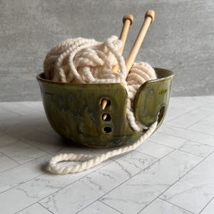 May include: A green ceramic yarn bowl with a cream-colored ball of yarn and two wooden knitting needles. The bowl has a curved design with holes for the yarn to pass through. The yarn is a thick, textured material.