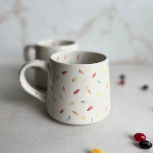 May include: Two white ceramic mugs with a confetti pattern in shades of pink, yellow, blue, and green. The mugs have a rounded shape and a handle.