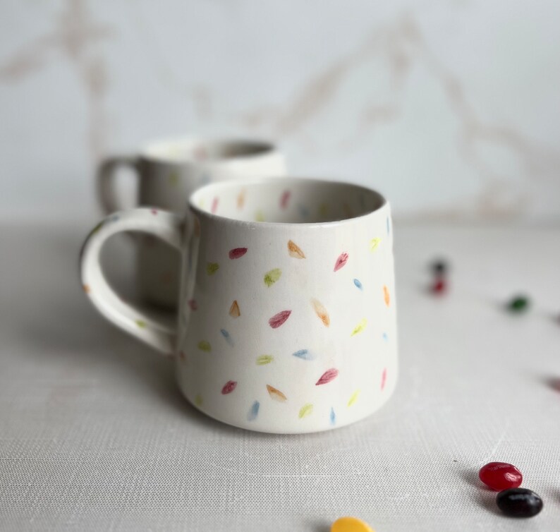 May include: Two white ceramic mugs with a confetti pattern in shades of pink, yellow, blue, and green. The mugs have a rounded shape and a handle.