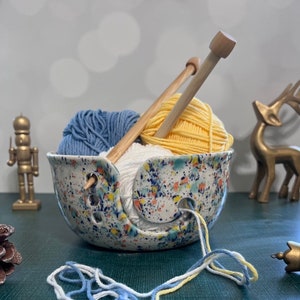 May include: A ceramic yarn bowl with a speckled white, blue, yellow, and green glaze. The bowl has three holes for yarn to feed through. Two wooden knitting needles are in the bowl with balls of blue, yellow, and white yarn.