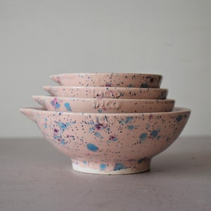 May include: A set of four pink ceramic measuring bowls stacked together. Each bowl has a spout and is speckled with blue and purple paint. The bowls are labeled with measurements.