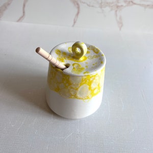May include: A white ceramic honey pot with a yellow glaze and a wooden honey dipper. The pot has a lid with a small loop handle.