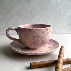 May include: A pink ceramic teacup and saucer set with a speckled design in blue and purple. The cup has a curved handle and sits on a matching saucer. Two brown, rolled wafer cookies are in the foreground.