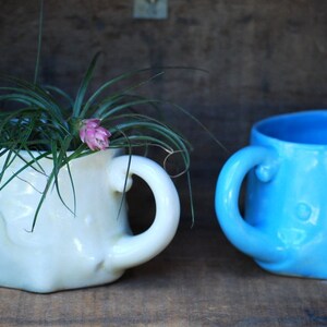 Puede incluir: Dos tazas de cer&aacute;mica con forma de elefante. Una es de color crema y contiene una planta con hojas verdes largas y una flor rosa. La otra taza es de color azul claro. Ambas tazas tienen un asa y est&aacute;n sobre una superficie de madera.