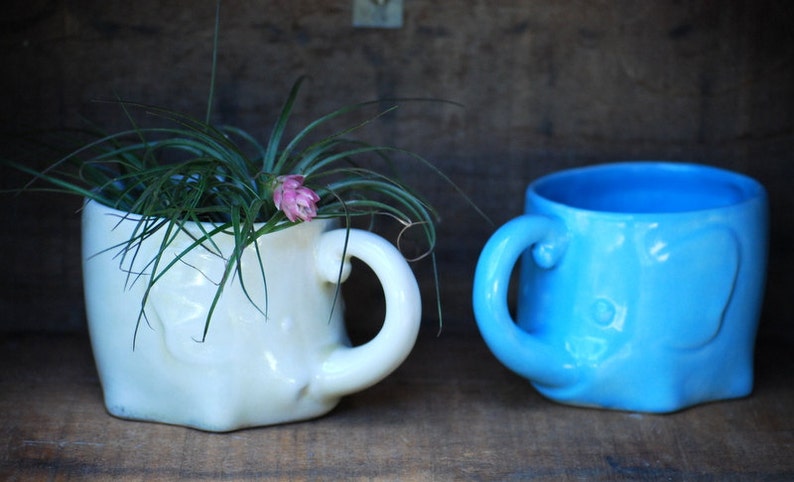 Puede incluir: Dos tazas de cer&aacute;mica con forma de elefante. Una es de color crema y contiene una planta con hojas verdes largas y una flor rosa. La otra taza es de color azul claro. Ambas tazas tienen un asa y est&aacute;n sobre una superficie de madera.