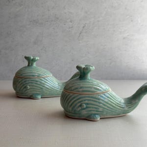 May include: Two ceramic whale-shaped containers with lids. The whales are a light blue-green color with white stripes. The containers are sitting on a white surface.