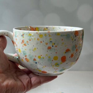 May include: A white ceramic teacup and saucer set with a speckled design in orange, yellow, blue, and green. The teacup has a large handle and a wide bowl. The saucer is visible at the bottom.
